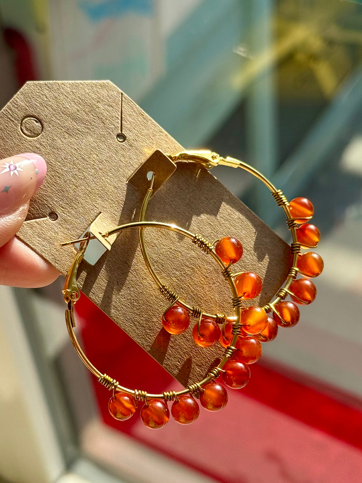 Gold hoop earrings with dangling amber-orange beads, displayed on a brown cardboard tag held by a hand.