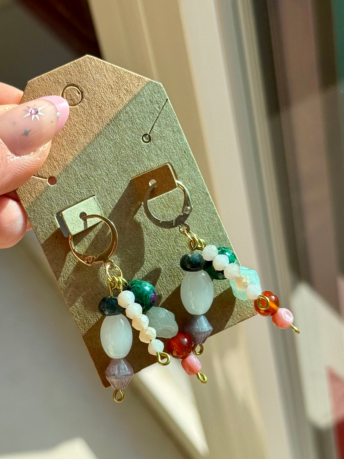 A pair of bohemian-style beaded dangle earrings on a kraft display card, held by a hand. The earrings feature gold-tone rings, white and light green beads, dark green beads, and small red and orange accents.